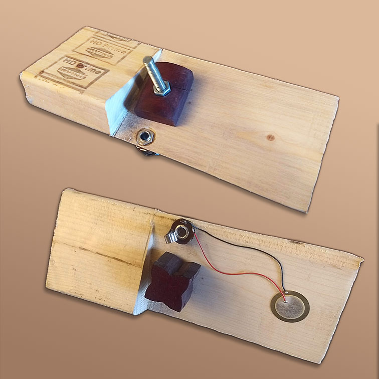 The back side of this daxophone clamps to a table and is clearly an unfinished 2x4. The rest of the daxophone has been cut at an angle to form a thin flat plate. Near the back side a rectangular clamp connects via a bolt through the flat plate to a wooden knob on the bottom. On the underside, a piezo element is wired to a quarter-inch jack that opens out to the top. 